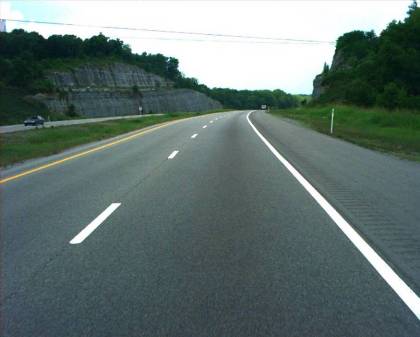 Figure 28.  Photograph.  SR-840.  The photograph shows the divided highway on State Route (SR)-840 near Nashville, TN, on which one of the pavement marking test decks was installed.  Each direction of travel has two lanes.  The photograph is taken from the driver's perspective from the right lane.  There is a yellow edge line on the left of the two lanes and a white edge line on the right of the two lanes.  Also, there are white dashed lines separating the two lanes.  The lanes in the opposing direction with one car are to the left, separated by a grassy median.