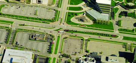 The photograph shows an aerial view of a restricted crossing U-turn (RCUT) intersection in Troy, MI. A tow lane minor road intersects a major six-lane road. At the intersection, the through movements for the minor road are blocked using a median. However, left-turn movements are allowed from the major road to the minor roads. Openings in the median are provided on either side of the intersection on the major road for allowing vehicles to make a U-turn. The through traffic from the minor street must make a right turn into the main street, make a U-turn in the median, and take a right turn to continue on the minor road. 