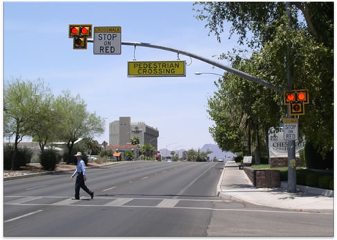 This photo shows a pedestrian walking to the left across a High-intensity Activated crossWalK (HAWK) at an intersection on an urban six-lane divided highway. A post at the right side of the road has a three-lens beacon head with one yellow lens centered beneath the two top red lenses. Both red lenses are illuminated. There is a sign below the beacon head that reads "CROSSWALK, STOP ON RED." The sign is rectangular and white, with "CROSSWALK" on a yellow background. A mast arm mounted on the post shows a similar beacon head with both red lenses illuminated and a similar sign, along with a hanging sign that reads "PEDESTRIAN CROSSING." The pavement markings are high-visibility, ladder-style crosswalk markings.