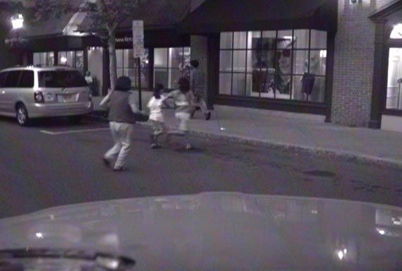 This photo shows an adult and two children crossing a road at midblock during the evening in front of a vehicle. There are stores on the side of the road as well as a parked vehicle.