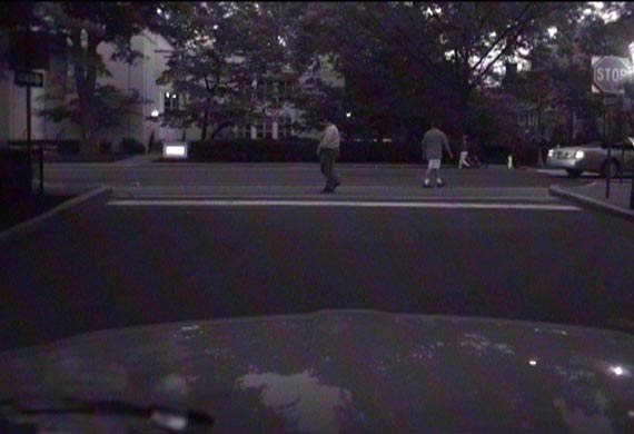 This photo shows pedestrians crossing a street at night in an urban environment at an unsignalized intersection in front of a vehicle. There are also pedestrians crossing the street away from the vehicle at midblock.