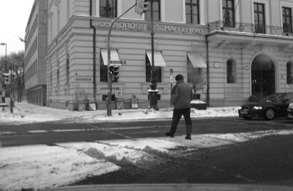 This photo shows a pedestrian crossing a snow-covered street at a signalized crosswalk in an urban environment while vehicles are waiting for the signal to change.