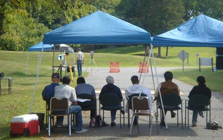 This photo shows the outdoor experimental setup from the rear of the participants’ tents at the Turner-Fairbank Highway Research Center (TFHRC). The photo shows the road on the research facility grounds where the experiment was conducted. In the background are grass and trees. The experimenter at his laptop workstation and a cooler for water are in the foreground. Two blue tents are side by side, with six research participants seated in a row beneath the tents. Further in the distance are the second experimenter’s table and tent along with the stimulus tripod and the spectroradiometer on its tripod. In the far distance, two orange traffic barriers isolate the experimental section of roadway from traffic.