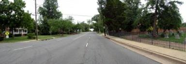 This photo shows 1 of 14 roadway panoramas presented to participants in the similarity rating task. A straight four-lane undivided road is shown, and there is no shoulder that would allow parking. A sidewalk abuts the curb on the right. A wrought-iron fence abuts the sidewalk. Gravestones are visible through the fence. On the left, the sidewalk has a small grass strip separating it from the curb, and a residence is visible. A pedestrian-crossing warning sign is visible on the left, although there is no marked crosswalk. There are mature trees on both sides of the road.
