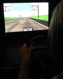 This photo shows an example of the roadway and sign view for the study. A participant is photographed from behind sitting in the simulator and looking at the screen.