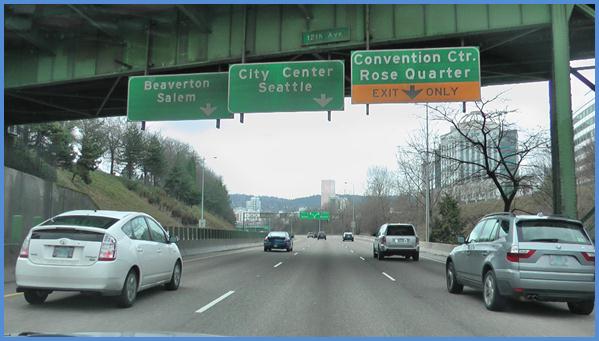 This photo shows the driving image for scenario 2 critical point 1. There is a three-lane single direction highway with cars traveling on it. There are three green signs: one above each of the lanes. The sign on the left has a down arrow and is labeled  Beaverton Salem.  The middle sign has a down arrow and is labeled  City Center Seattle.  The sign on the right is green on the top half and is labeled  Convention Ctr. Rose Quarter.  The bottom half is yellow with a down arrow and is labeled Exit Only. 