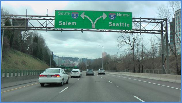 This photo shows the driving image for scenario 2 critical point 2. There is a three-lane single direction highway with cars traveling on it. There is a green sign that spans all three lanes. It shows an illustration of the highway with three lanes that splits two lanes to the left and two lanes to the right. The middle lane is an option lane. The left side is labeled  5 South Salem,  and the right side is labeled  5 North Seattle. 