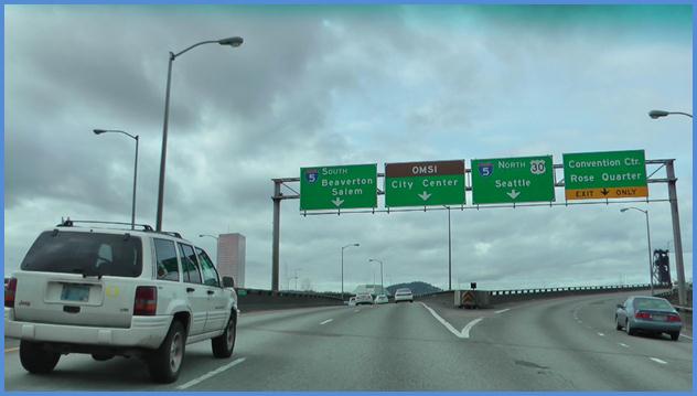 This photo shows a driving image for scenario 2 critical point 4. There is a three-lane single direction highway with vehicles traveling on it. The lanes are shown splitting to the left and right. The middle lane is acting as an option lane, where drivers can choose to drive to the left or to the right. There are two lanes to the left and two lanes to the right. There is a green sign above each of the four lanes. The leftmost sign has a down arrow and is labeled  5 South Beaverton Salem.  The next sign also has a down arrow. The top half of the sign is brown and is labeled  OMSI.  The bottom half is green and is labeled  City Center.  The third sign has a down arrow and is labeled  5 North 30 Seattle.  Finally, the right-most sign has a down arrow. The top part of the sign is green and is labeled  Convention Ctr. Rose Quarter.  The bottom part is yellow and is labeled as an exit only lane.