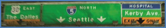this photo shows guide signs associated with scenario 3 critical point 1. There are three signs next to each other. For the sign on the left, the top half is green and is labeled  30 East The Dalles.  The bottom half is yellow with a down arrow and is labeled  Exit Only.  The middle sign is green with two down arrows and is labeled  5 North Seattle.  The right sign is blue on the top and is labeled  Hospital.  The middle part is green and is labeled  Kerby Ave.  The bottom part is yellow with a down arrow and is labeled  Exit Only. 
