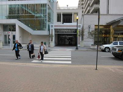 Figure 19. Photo. Example of pedestrians outside of crosswalk at site on 23rd Street. Unsignalized pedestrian crosswalk near Crystal City Mall in Arlington, VA. The midblock crosswalk is marked with a ladder design and crosses a one-way roadway with two through lanes and parking on the right side of the road. Three pedestrians are crossing the roadway—one inside the marked crosswalk and two just outside of the marked crosswalk. There is a car yielding in advance of the crosswalk a pedestrian crosswalk sign (W11-2) at either end of the crosswalk.