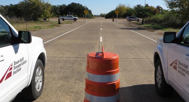 Figure 4. Photo. View of the study assemblies. This photo shows a view of the study site crosswalk from the subject drivers' point of view. In the foreground, there is an orange work zone barrel in the center of the two-lane road, and cars are parked on either side of the barrel. In the background is the study site crosswalk, with a pedestrian photograph cardboard cutout on the right side of the road at the crosswalk and a rectangular rapid flashing beacon and sign on either side of the road, which are attached to metal poles. The rectangular flashing beacons are flashing.