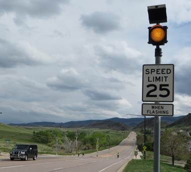 This photo shows a road next to a sign assembly that has (starting from top) a solar panel, a 12-inch yellow circular beacon, a black-on-white sign that says “SPEED LIMIT 25,” and a black-on-white plaque that says “WHEN FLASHING.” In the background is a pedestrian crossing. 