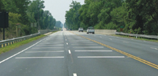 This is a photo of a four-lane road, two lanes of traffic per side. There are white transverse pavement markings painted onto the travel lanes that span almost the entire width of the travel lane.