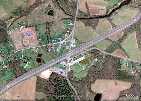 This photo shows a satellite view of the restricted crossing U-turn (RCUT) intersection that was the subject of this study. A four-lane highway, US-15, is shown extending from the bottom left to the top right. A two-lane rural road, US-15 Business, intersects the highway in the north-south direction. The main intersection has an opening that allows traffic on the divided highway to turn left onto the local road. Curbing can be seen that blocks traffic from the minor road from turning left through the opening at the main intersection. Local road traffic must turn right at the main intersection. Directional U-turn openings can be seen in the highway median 1,800 ft on either side of the main intersection. These openings allow local road traffic to make a U-turn to complete through or left-turn movements.