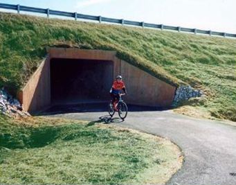 2) An underpass continues this shared use bicycle path beneath a four-lane highway with high traffic volume. In this photo, a child rides a bike through a wide underpass that leads to a trail. The road over the underpass is a four-lane highway with heavy traffic.