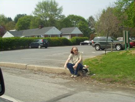 The first picture shows a woman sitting on the curb of the road waiting for a bus. There is no sidewalk, only a paved shoulder. 