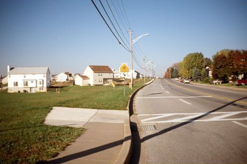Picture shows a very wide suburban street with the backs of the neighborhood houses facing the street. In the foreground of the picture, there is a sidewalk with a mid-block crossing that ends short or reaching the neighborhood.
