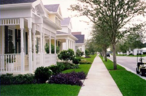 Picture shows classic American homes with large front porches. In front of the homes is a tree lined sidewalk as a buffer between the homes and the street. Driveways and garages are not visible.