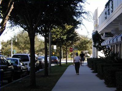 The first picture shows two pedestrians walking on a sidewalk in front of stores. The sidewalk is wide with a grass strip planted with trees between the sidewalk and street. There are cars parked along the street. 