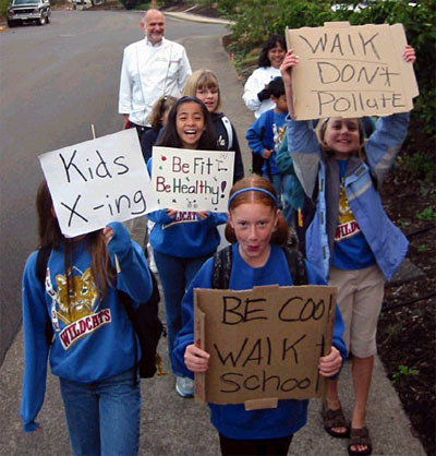 The picture shows 6 children holding up handmade signs with messages such as 'Kids Crossing', 'Be fit - Be healthy', 'Walk don't pollute' and 'Be cool, walk to school.'