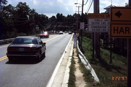 First picture shows a road with trees on one side and utility poles and guard rail on the other side. Pedestrians have worn a dirt path into the one foot wide strip between the guard rail that runs in front of the utility poles and the curb. A pedestrian is shown walking precariously on the path.