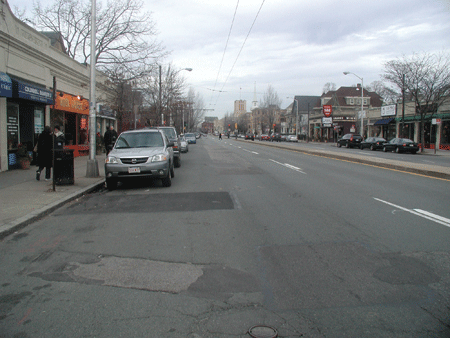 Figure 2. Photo. Massachusetts Avenue condition in the before period. The photo shows Massachusetts Avenue in Cambridge, MA, in the before sharrow condition. The street is a two-way street, with two travel lanes in each direction and a raised median in the center. The photo is shot from the left side of the street with parked cars on the left side facing forward, and there are stores along both sides of the street.