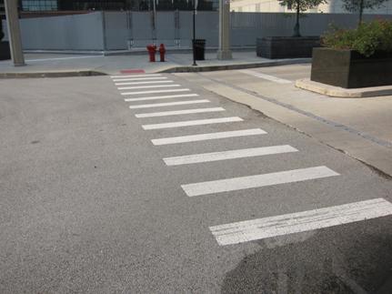 This photograph shows continental markings across a street as viewed from the curb. The markings consist of several parallel, white vertical bars across the width of the street.