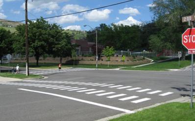 This photograph shows double continental markings as viewed from the roadside. The markings consist of two sets of parallel, white vertical bars running across the width of the street. The sets of bars form two horizontal rows with about the width of the sidewalk between them. 