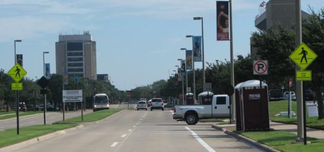 This photograph shows the distance view of an existing midblock site with transverse marking before the marking was repainted. The crosswalk spans two lanes of a four-lane campus street at Texas A&M University. On roadside poles to the left and right of the street and slightly ahead of the crosswalk are yellow pedestrian warning signs (W11-2).