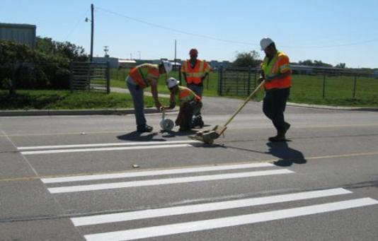 This photograph showing workers installing markings used at one of the study sites. There are four men wearing orange traffic safety vests. The men are putting down white marking tape to construct a bar-pairs style crosswalk.