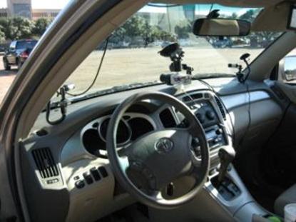 This photograph shows the interior of the instrumented vehicle used in the study. Three cameras are positioned to face out the front windshield. The largest one is in the center of the SUV, fixed atop the dashboard near the center console. It records the forward view. The other two cameras are attached to the windshield on the right and left sides of the SUV and are angled to record the side views.