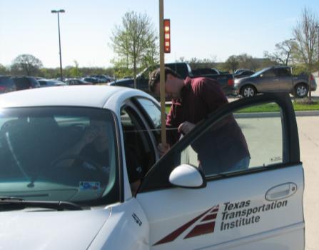 This photograph shows the method used for measuring driver eye height. A participant is seated in driver's seat of the vehicle, holding a string at his nose. An experimenter is standing outside the open door of the car, holding the scale upright on the ground. The experimenter is holding the other end of the string and is using it to read the participant's eye height.