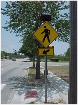 A pole mounted at the side of a roadway near a crosswalk bears the W11-2 pedestrian warning sign, which shows a silhouette of a person walking to the left. The sign is a yellow diamond with a black border, and the silhouette is black. Below the sign, there is a rectangular rapid-flashing beacon (RRFB) with two yellow lights, and the light on the right is activated. Below the sign, there is a rectangular yellow sign with a black border and a black arrow pointing diagonally downward to the left toward the crosswalk. Above the W11-2 sign, there is a photovoltaic panel.