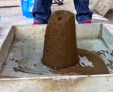 Photograph of a slump test specimen performed on stiff, low-slump, freshly mixed concrete made with alternative calcium aluminate cement. The slump specimen is in a metal mixing pan. It is a dark brown color, and the slump specimen is standing upright.