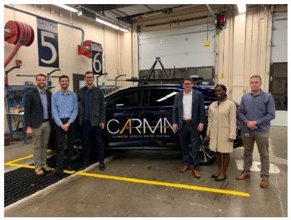 Three people -- 2 men and 1 woman -- stand on the right of the photo. Another set of three people -- three men -- stands on the left of the photo. Both teams stand in front of a CARMA vehicle in the Saxton Transportation Operations Laboratory at Turner-Fairbank Highway Research Center.