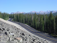 Beartooth Highway curves at the base of a rock slope with evergreen forests and rugged mountains in the background. In the early 1930’s, Beartooth Highway was constructed as a long approach road to Yellowstone.