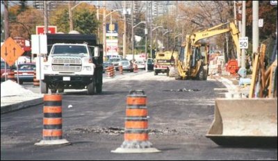 A street next to a roadway is blocked off as a construction site, thus causing congestion on the adjacent roadway. A dump truck, hoe ram, and front end loaders are on site.