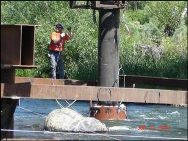 A construction worker stands near an underwater pile driving hammer with an operating bubble curtain surrounding the pile and hammer. The bubbles are visible at a few feet higher than the water surface at the top of the curtain.