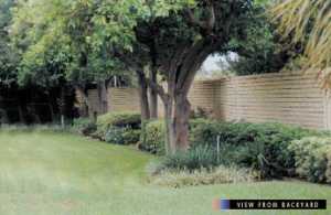 photo:  precast concrete noise barrier viewed from the backyard of a home, with a brick-look barrier surface and existing trees and abundant vegetation along the barrier (photo is intended to illustrate a typical example of a pleasant backyard view of a well-designed and well-constructed noise barrier)