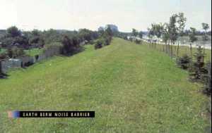 photo: viewed from the top of an earth berm, showing trees planted on the berm, the backyards of homes, and a highway with traffic (photo is intended to illustrate a typical example of a well-designed and well-constructed earth berm noise barrier)