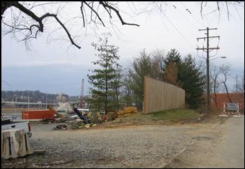 A storage area for supplies and equipment (as noted by the crane) in a residential location that is placed far from residences and near the highway right-of-way. A temporary wooden barrier can also be seen partially shielding the storage area.