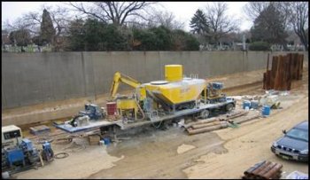 A construction site in the highway right-of-way uses an existing retaining wall to act as a noise barrier for a stationary grout injection activity. An historic cemetery and residences are in the background. 