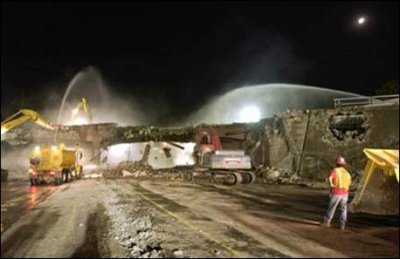 A nighttime demolition of an existing concrete bridge span has a loader, bulldozer, and several water sprayers to minimize dust.