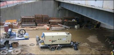 An equipment and storage yard, noted by a compressor and steel pipes, are located near an adjacent retaining wall and underneath an existing bridge span. 