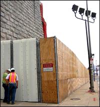 An existing structure, 4 stories tall, and plywood on chain link fence along with a gate and absorptive mat combination shield a construction site.