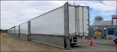 Six tractor trailers used for storage are aligned as a barrier to shield adjacent residences and businesses from a construction site. Vinyl sheeting extends from the back and bottom of the trailers to completely shield the line-of-sight to the residences.