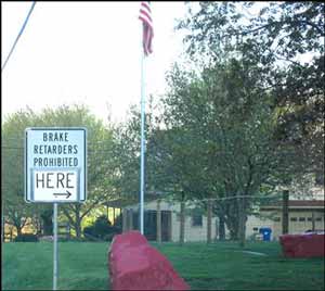 An informational street sign (white background with black letters) declaring brake retarders (engine compression brakes) are prohibited, with modification of the sign adding a directional arrow and the words “here”.