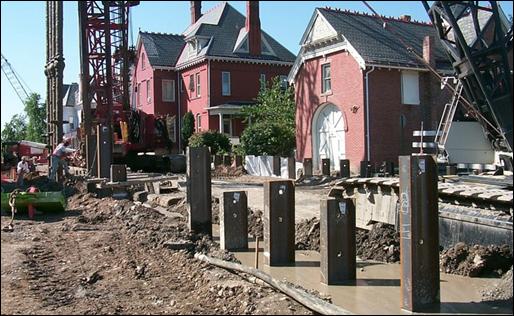 A crane and pile driving equipment operate within 25 feet of nearby residences. Many piles can be seen driven into the soil.