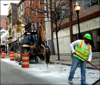 A backhoe loader with jackhammer attachment is smashing pavement in a downtown, urban setting. 