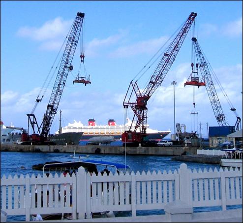 Several cranes working near a boardwalk where a cruise ship passes by in the background.